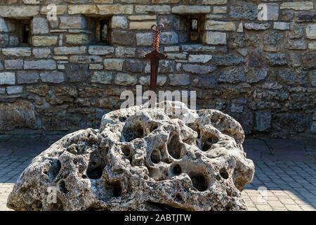 Ein altes Schwert in einen großen Stein gegen eine Mauer aus Stein mit kleinen Fenstern. Stockfoto