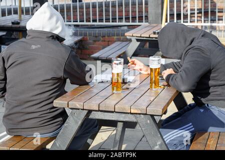 Zwei männliche Jugendliche sitzen an einem Picknicktisch vor einem Pub, tragen dunkle Kapuzenpullover, sind mit Gesicht bedeckt und rauchen und trinken Bier. VEREINIGTES KÖNIGREICH Stockfoto