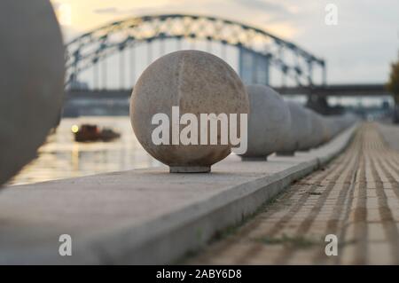 Nahaufnahme von Steinkugeln dekorieren Daugava Promenade in der Perspektive, mit dem Boot am Fluss Daugava und Eisenbahnbrücke in Sonnenuntergang Licht im Hintergrund Stockfoto