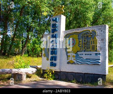 Ein Schild mit den Namen von Tschernobyl in der Ukraine geschrieben. Oben auf dem Denkmal ist eine gelbe Atom. Auf der Seite gibt es einen industriellen Fabrik. Cher Stockfoto