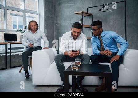 Das Sitzen auf einer Couch, trinkt Whiskey und haben diskutiert. Gruppe der multirassischen Büroangestellte in formelle Kleidung sprechen über Aufgaben und Pläne Stockfoto