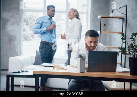 Lächelnd Mitarbeiter und Hintergrund. Junge Unternehmer sitzt auf dem Stuhl vor Laptop und arbeitet mit Berichten Stockfoto