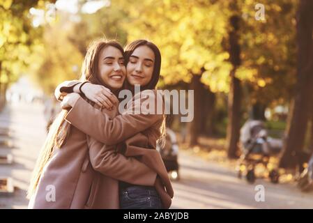 Seite Profil der junge lächelnde brunette Doppel Mädchen umarmt und Spaß in lässig-Mantel an Herbst sonnigen Park Alley im Stehen, in der Nähe von einander Stockfoto