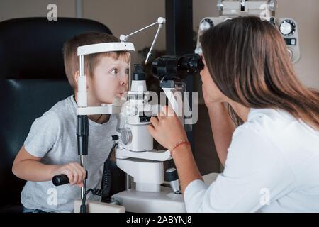 Die lustige Gesichter. Little Boy In Test für seine Augen mit speziellen optischen Apparat von weiblichen Arzt Stockfoto