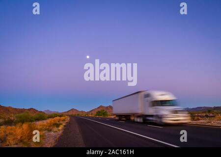Gezielt verwischt 18 rad Fernverkehr Lkw auf der Autobahn in der Wüste bei lila und blau Sonnenaufgang mit Mond im Himmel Stockfoto