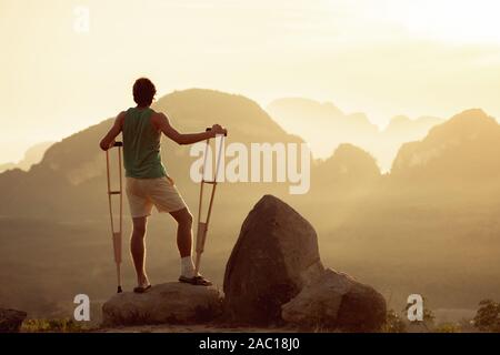 Verletzte Mann steht mit Krücken auf Big Rock. Reisen für behinderte Menschen Konzept Stockfoto