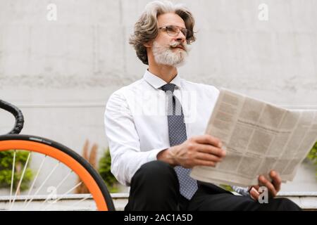 Foto der Grauhaarige bärtigen Geschäftsmann in Brillen lesen Zeitung während sitzt auf der Bank mit dem Fahrrad im Freien Stockfoto
