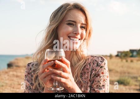 Foto fröhliche blonde Frau hält Glas Wein und lächelnd beim Spaziergang in der Nähe von Seaside während der sonnigen Tag Stockfoto