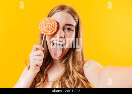 Bild von einer fröhlichen, glücklichen jungen niedlich Jugendmädchen über gelb Wand Hintergrund holding Süßigkeiten Lutscher nehmen durch die Kamera posiert selfie isoliert Stockfoto