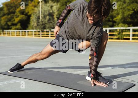 Foto von sportlichen jungen Mann in Sportkleidung Übungen auf der Matte, während sie auf dem Spielplatz im Freien arbeiten Stockfoto