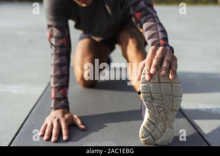 7/8-Foto von athletischen Mann in Sportswear stretching sein Bein auf der Matte, während sie auf dem Spielplatz im Freien arbeiten Stockfoto