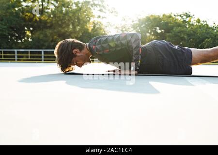 Foto von athletischen maskulinen Mann in Sportswear Übungen auf der Matte, während sie auf dem Spielplatz im Freien arbeiten Stockfoto