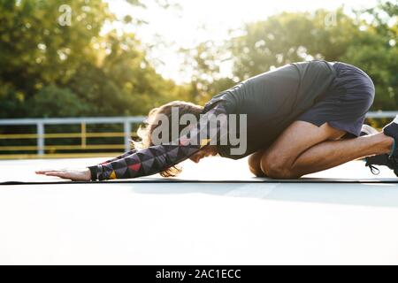 Foto von Konzentriertem brünett Mann in Sportswear Übungen auf der Matte, während sie auf dem Spielplatz im Freien arbeiten Stockfoto