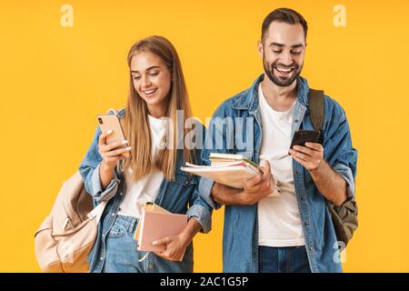 Porträt eines attraktiven fröhlicher junger Studenten Paar Tragen von Rucksäcken über Gelb Hintergrund stehend, mit Handys, holding Buch Stockfoto