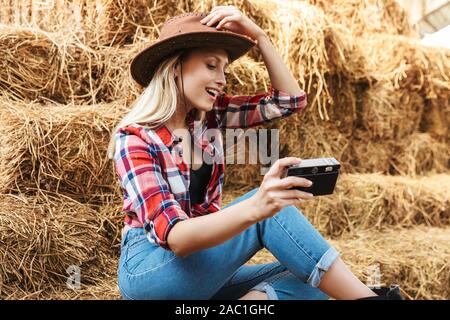 Lächelnde junge blonde Cowgirl sitzt auf einem Heuhaufen an der Scheune, Aufnehmen von Bildern mit der Kamera Stockfoto