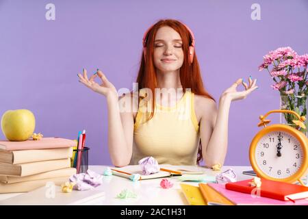 Foto von entspannt Schüler Mädchen in Kopfhörer am Schreibtisch sitzend mit Übung Bücher während der Hausaufgaben über lila Hintergrund isoliert Stockfoto