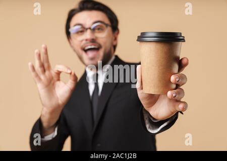 Bild der kaukasischen Geschäftsmann in Anzug, ok Zeichen und Holding takeaway Kaffeetasse isoliert über beigen Hintergrund Stockfoto