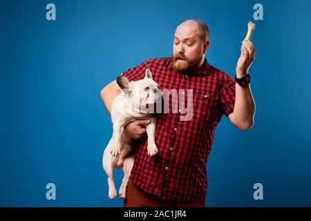 Mann im roten Hemd hält Hund und Knochen auf leeren blauen Hintergrund im Studio Stockfoto