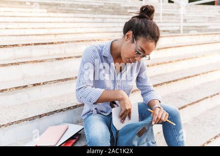 Attraktive junge afrikanische Frau legere Kleidung sitzen auf Schritte im Freien, unter Bücher aus ihrem Rucksack Stockfoto