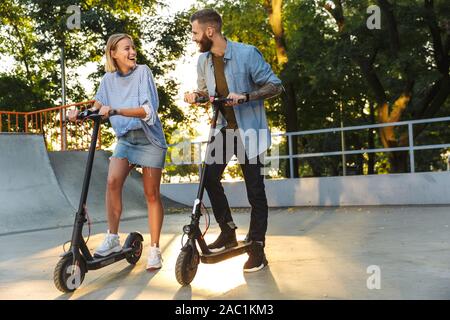 Image der Jungen glückliches Paar in Freizeitkleidung lächelnd gekleidet beim Reiten e-Scooter zusammen am Sportplatz Stockfoto