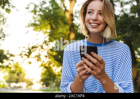 Bild des Blonden fröhliche Frau in Freizeitkleidung mit Handy und Ohrhörer gekleidet, während er im Green Park Stockfoto