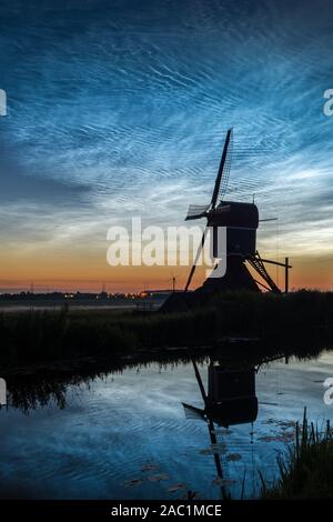 Silhouette einer alten holländischen Windmühle gegen einen Himmel mit leuchtenden Nachtwolken (NLC) um Mitternacht Stockfoto