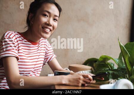 Bild einer positiven junge schöne asiatische Frau sitzen im Cafe mit Handy, Kamera. Stockfoto