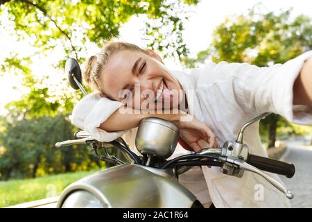 Schönen lächelnden jungen Mädchen legere Sommerkleidung sitzen auf einem Roller draußen auf den Straßen der Stadt, die eine selfie Stockfoto