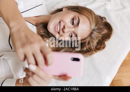 Bild des lächelnden Jungen Mädchen mit weißen Pyjama holding Handy während in Bett zu Hause liegen Stockfoto