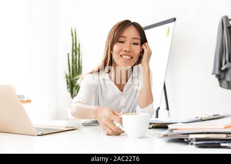 Lächelnd asiatischen Geschäftsfrau Musikhören mit Kopfhörern beim Trinken Kaffee im Büro Stockfoto