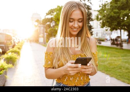 Schöne junge blonde Mädchen mit Handy beim Stehen im Freien Stockfoto