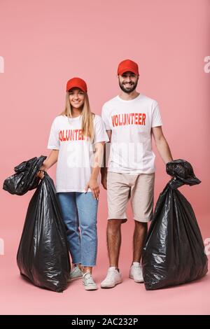 Die volle Länge eines glücklichen jungen Freiwilligen Paar stehend über rosa Hintergrund isoliert, holding Müllsäcke Stockfoto