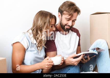 Foto von lustig Paar Kaffee trinken und lesen Magazin beim Sitzen in der Nähe von Kartons über weiße Wand isoliert Stockfoto