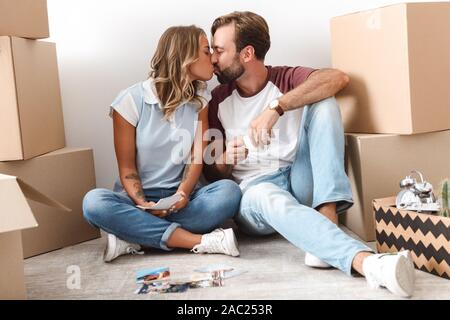 Foto von romantisches Paar legere Kleidung, trinken Kaffee und Küssen beim Sitzen in der Nähe von Kartons über weiße Wand isoliert Stockfoto