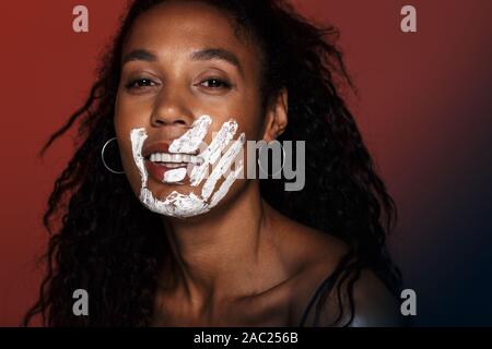 Nahaufnahme eines lächelnden jungen afrikanischen Frau mit weißer Hand Druck auf ihr Gesicht über den roten Hintergrund isoliert Stockfoto