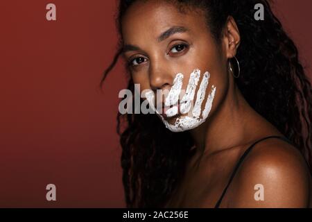 Nahaufnahme eines jungen afrikanischen Frau mit weißen Hand Druck auf ihr Gesicht über den roten Hintergrund isoliert Stockfoto