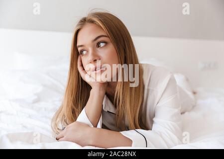 Foto der hübschen blonden Frau im Pyjama zur Seite schauen, während Sie auf dem Bett nach dem Ruhezustand oder nap liegen Stockfoto