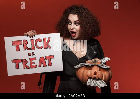 Bild von Angeregten Hexe girl Holding Halloween Kürbis und Trick oder Festlichkeit über rote Wand isoliert Stockfoto