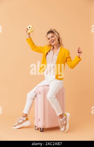 Bild von fröhlichen freuen positive Frau Touristen sitzen auf Koffer über beige Wand Hintergrund holding Kamera isoliert. Stockfoto