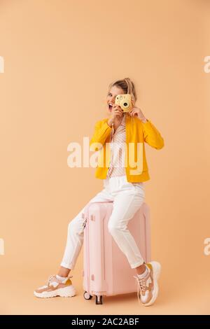 Bild von optimistisch gerne Frau Touristen sitzen auf Koffer über beige Wand Hintergrund holding Kamera isoliert. Stockfoto