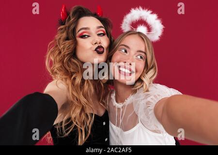 Foto von einem positiven süße Frauen Engel und Dämon in der Karneval Kostüme über rote Wand Hintergrund nehmen ein selfie durch die Kamera küsst weht. Stockfoto