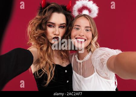 Foto eines Glücklich lächelnde niedliche Frauen Engel und unverschämt Daemon in Karneval Kostüme über rote Wand Hintergrund nehmen ein selfie durch die Kamera. Stockfoto