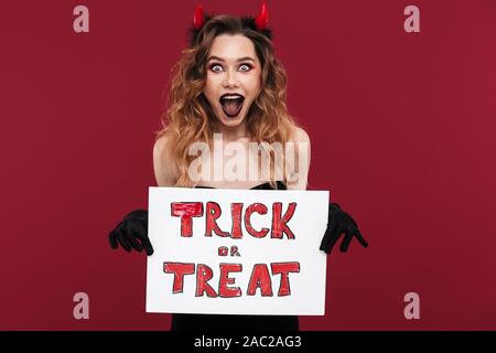 Bild von Schockiert Frau Daemon in Karneval Kostüm über rote Wand Hintergrund Holding copyspace leer mit Halloween Trick oder Text behandeln isoliert. Stockfoto