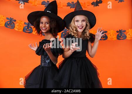 Bild von Nizza multinationale Mädchen in Schwarz halloween Kostüme lächelt und macht sich lustig über orange Kürbis Wand isoliert Stockfoto