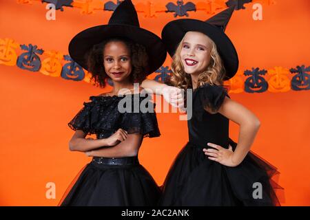 Bild von niedlichen multinationale Mädchen in Schwarz halloween Kostüme lächelnd und ständigen zusammen über orange Kürbis Wand isoliert Stockfoto