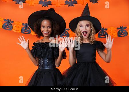 Bild von Angeregten multinationale Mädchen in Schwarz halloween Kostüme lächelnd mit wirft Hände isoliert über orange Kürbis Wand Stockfoto