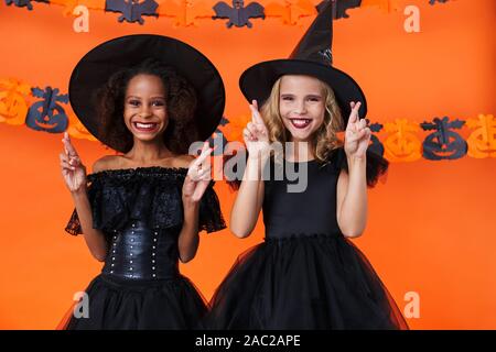 Bild des freudigen multinationale Mädchen in Schwarz halloween Kostüme lächelnd mit gekreuzten Fingern über orange Kürbis Wand isoliert Stockfoto