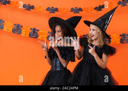 Bild von lächelnden multinationale Mädchen in Schwarz halloween Kostüme umarmen und Pointing Finger zur Seite über orange Kürbis Wand isoliert Stockfoto