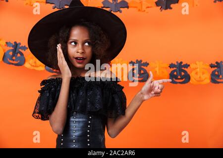 Bild von Angeregten African American Girl in Schwarz halloween kostüm Zeigefinger beiseite über orange Kürbis Wand isoliert Stockfoto