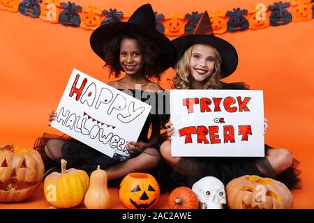 Bild von lächelnden multinationale Mädchen in Schwarz halloween Kostüme placards halten Sie beim Sitzen auf dem Boden isoliert über orange Kürbis Wand Stockfoto